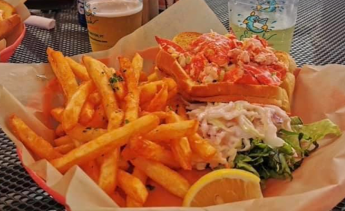 A plate of lobster roll with fries, coleslaw, lettuce, and a lemon wedge, served on a table with drinks in the background.