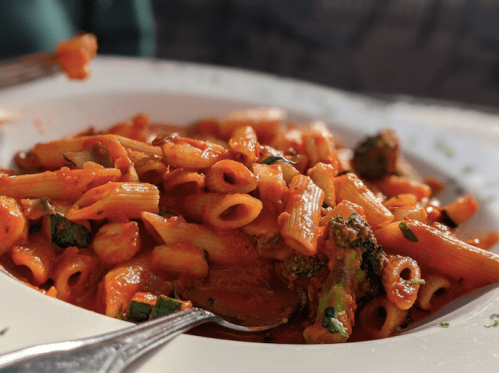 A close-up of a plate of pasta with tomato sauce, broccoli, and small pieces of meat, garnished with herbs.
