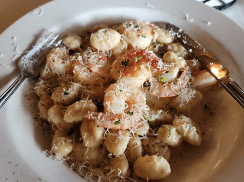 A plate of creamy gnocchi topped with shrimp and grated cheese, garnished with herbs and served with a fork and knife.