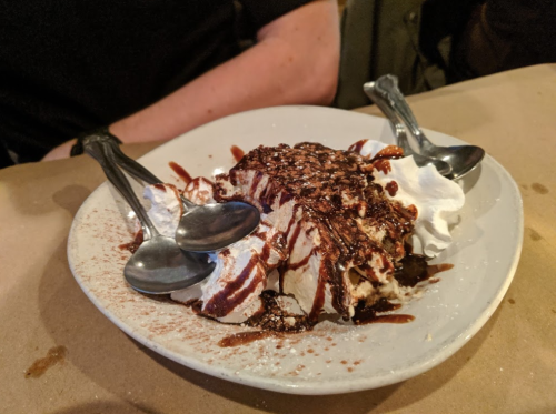 A dessert plate with whipped cream, chocolate sauce, and multiple spoons, set on a brown paper table covering.
