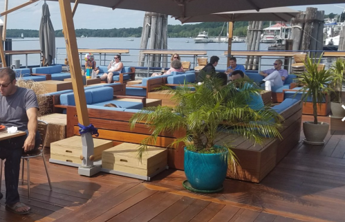 Outdoor seating area by the water, featuring wooden furniture, potted plants, and people relaxing on a sunny day.