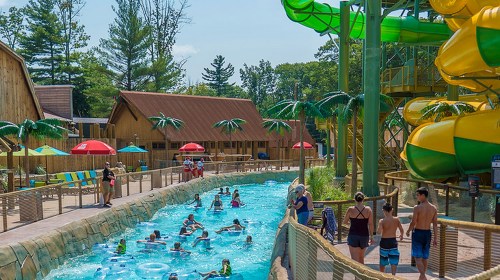 A water park scene with a lazy river, colorful slides, and people enjoying the sun near palm trees and cabanas.