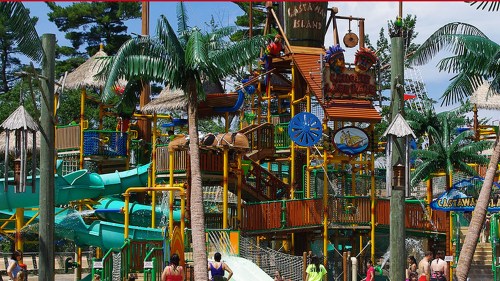 Colorful water park with slides, palm trees, and a large play structure, bustling with visitors enjoying the attractions.