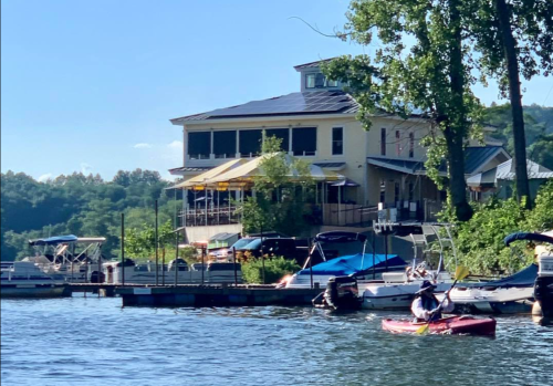 A person kayaking on a lake near a riverside restaurant surrounded by trees and boats.