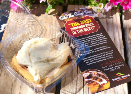 A cinnamon roll with cream cheese frosting in a takeout container, next to a promotional flyer for Parowan Visitor Center.