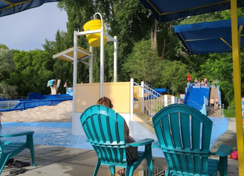 A water park scene featuring blue chairs, a splash pad, and slides in the background surrounded by greenery.
