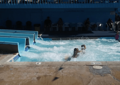 A water park scene with a wave pool, featuring people enjoying the water and slides in the background.