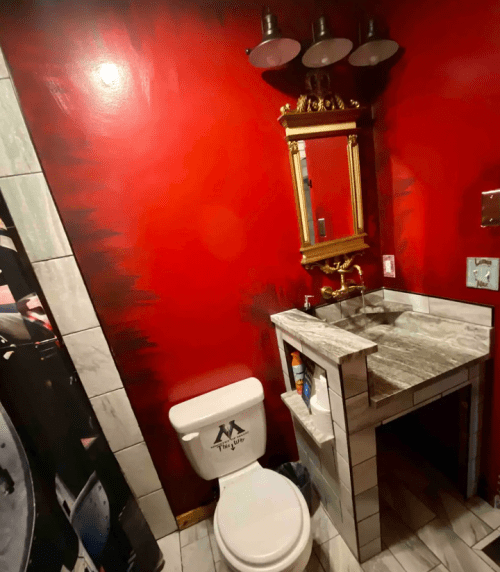A small bathroom featuring a red wall, a toilet, a marble sink, and a decorative mirror with lights above.
