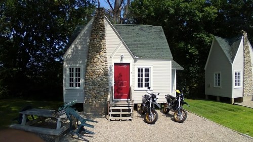 A small cottage with a red door and stone chimney, flanked by two motorcycles, surrounded by greenery.