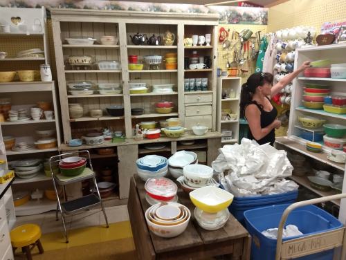 A woman organizes colorful bowls and dishes on shelves in a vintage shop, surrounded by various kitchenware.