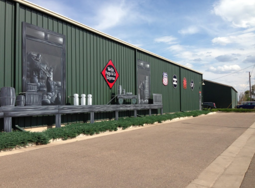 A green building with a mural depicting shelves and vintage items, under a blue sky with scattered clouds.