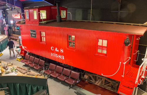 A large red caboose displayed indoors, with seating in front and various model trains in the background.