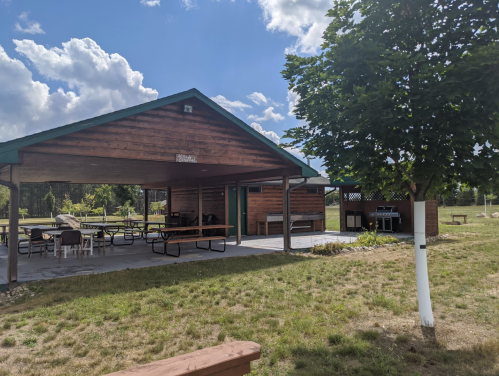 A wooden pavilion with picnic tables, surrounded by grass and trees, under a partly cloudy sky.