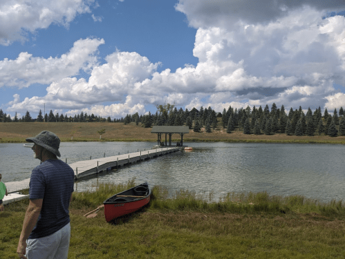 A man in a hat stands by a calm lake with a dock, canoe, and trees under a partly cloudy sky.