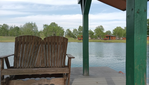 A wooden chair on a dock overlooking a calm lake, surrounded by trees and a clear sky.