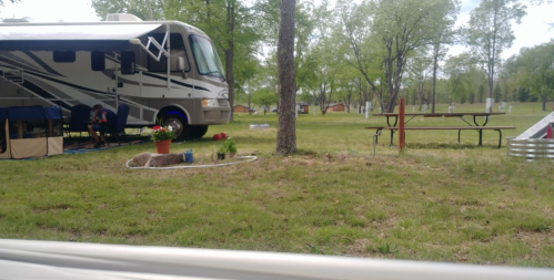 An RV parked in a grassy area with trees, a picnic table, and a small garden. A person is sitting nearby.