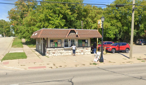 A small stone building with a sloped roof, surrounded by trees, and two people at a service window. Cars parked nearby.