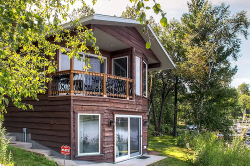 A cozy wooden cabin with a balcony, surrounded by trees and greenery near a lake.