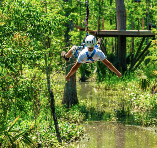 A person zip-lining over a lush green landscape with trees and a small body of water below.