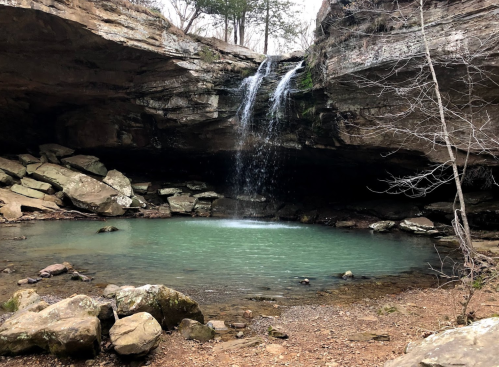 A serene waterfall cascades into a clear pool surrounded by rocky terrain and sparse trees.