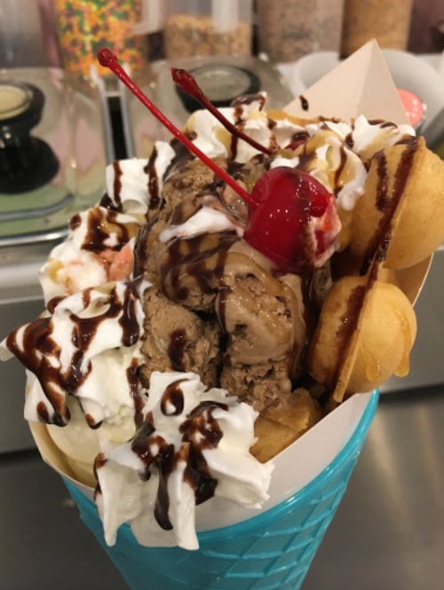 A colorful ice cream sundae in a blue cone, topped with whipped cream, chocolate sauce, cherries, and mini waffles.