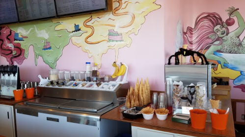 Colorful ice cream shop counter with toppings, cones, and a vibrant mural on the wall.