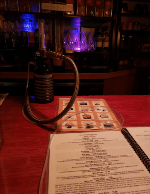 A bar counter with a menu and a unique drink dispenser in the foreground, colorful lights in the background.