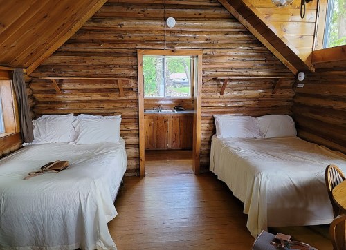 Cozy cabin interior with two beds, wooden walls, and a small kitchenette visible through an open doorway.