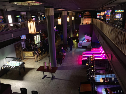 A dimly lit arcade with various games, including foosball and neon-lit machines, and people enjoying the space.