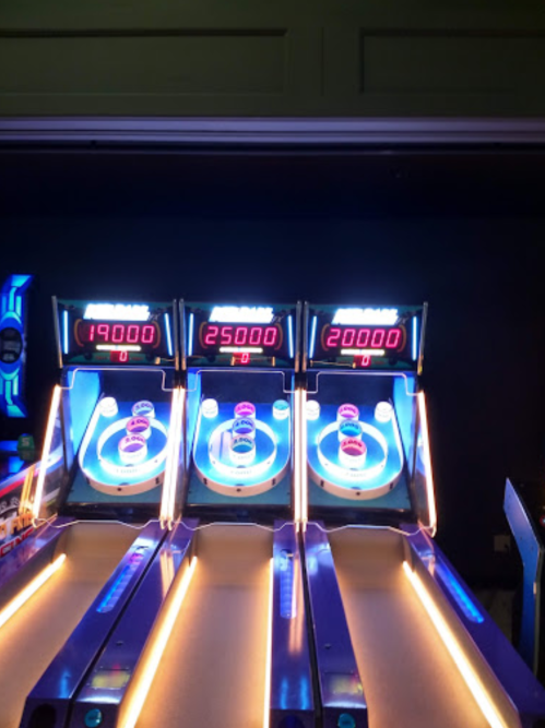 Three arcade skee-ball machines displaying scores of 19000, 25000, and 20000, with colorful lights and screens.