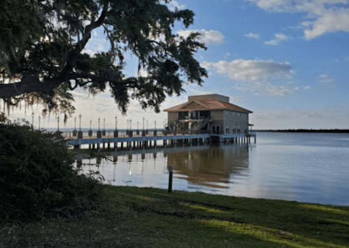 A waterfront building with a pier, surrounded by trees and reflecting in calm water under a blue sky.