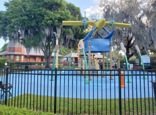 A colorful water play area with spray features, surrounded by a black fence and lush greenery.