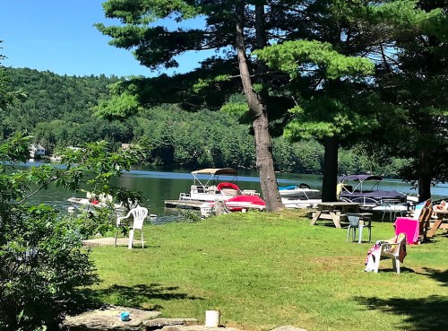 A serene lakeside scene with boats docked, green trees, and chairs on the grass under a clear blue sky.