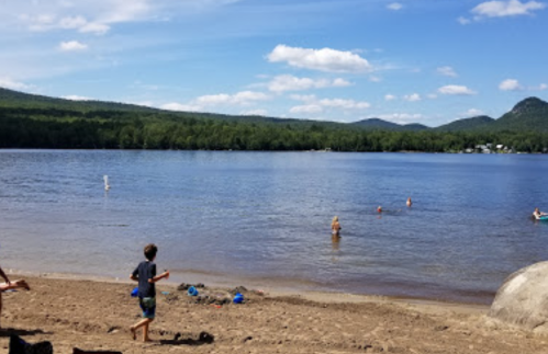 A sandy beach by a lake with people swimming and playing, surrounded by green hills and a blue sky with clouds.