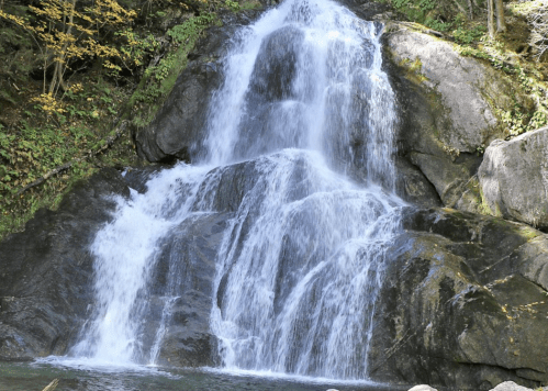 A cascading waterfall flows over large rocks, surrounded by lush greenery and autumn foliage.