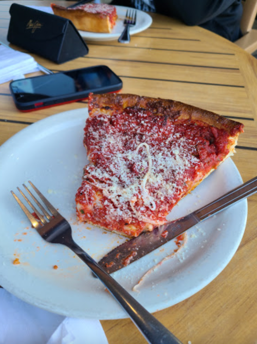 A slice of deep-dish pizza topped with marinara sauce and cheese on a plate, with utensils and a phone nearby.