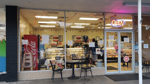 A bakery storefront with an "OPEN" sign, displaying pastries and a Coca-Cola vending machine outside.