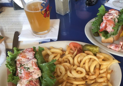 A plate with a lobster roll on lettuce, curly fries, and a glass of beer on a blue table.