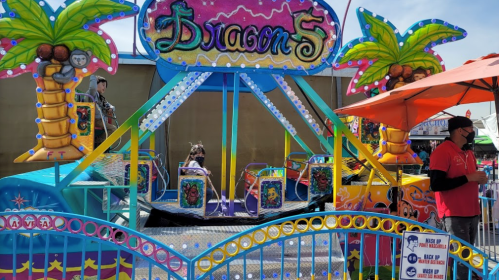 Colorful carnival ride named "Dragon 5" with palm trees, surrounded by a festive atmosphere and a person in a red shirt.