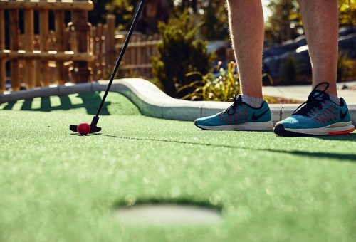 A person stands on a mini-golf course, preparing to putt a red ball with a black putter.