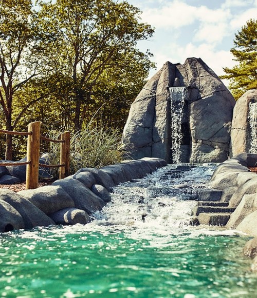 A serene water feature with a waterfall cascading over rocks into a turquoise pool, surrounded by trees and greenery.