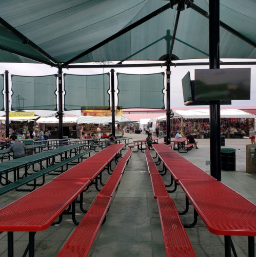 Empty red picnic tables under a shaded canopy, with a view of a bustling market in the background.