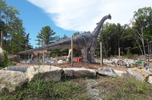 A large dinosaur statue stands among rocks and trees in a park, with visitors in the background.