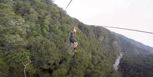A person zip-lining over a lush green forest with a river visible below.