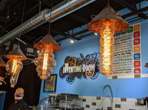 Three unique pendant lights hang from the ceiling in a colorful cafe, with a man in a mask standing nearby.