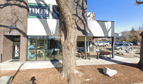 A storefront with large windows, a tree in front, and a parking lot visible in the background under a clear blue sky.