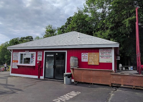 A red arcade building with a gray roof, featuring windows, a door, and a parking area in front. Trees in the background.