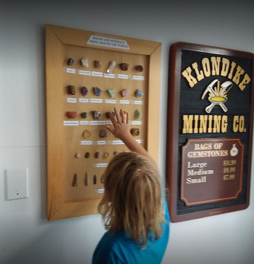 A child examines a display of various rocks and minerals at a mining exhibit, with a sign for Klondike Mining Co. nearby.