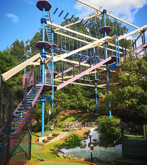Colorful ropes course structure with stairs, surrounded by greenery and a small waterfall in a recreational area.
