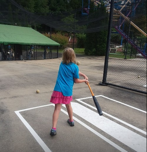 A young girl in a blue shirt and pink skirt swings a bat at a baseball in a batting cage.
